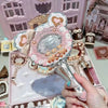 A hand holds a decorative makeup mirror set featuring pink and gold accents on a light pink background.