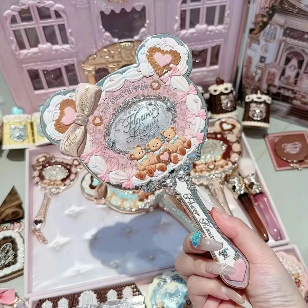 A hand holds a decorative makeup mirror set featuring pink and gold accents on a light pink background.