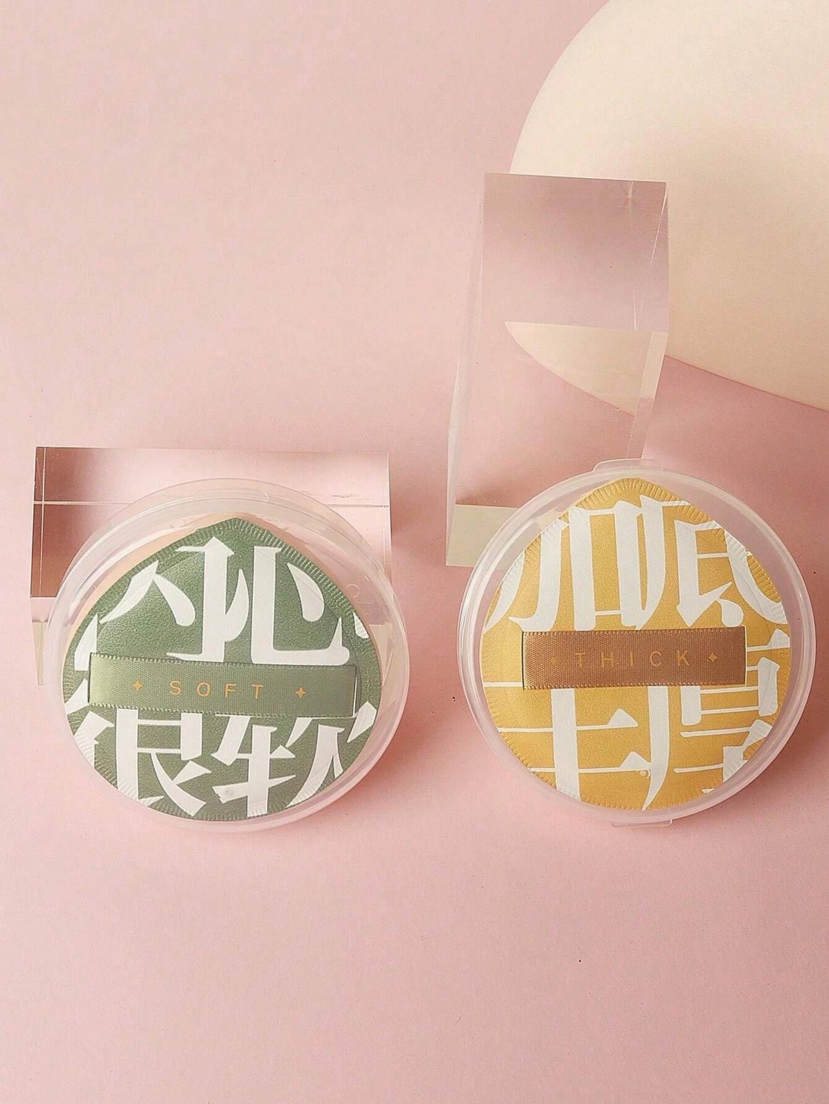 A pair of dual-sided makeup sponge puffs in green and yellow are displayed on a pink surface.
