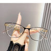 Une main tient une paire de lunettes de lecture élégantes à monture chat-œil transparentes avec des accents dorés sur un fond