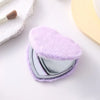 A heart-shaped foldable travel makeup mirror covered in fluffy purple faux fur resting on a white surface.