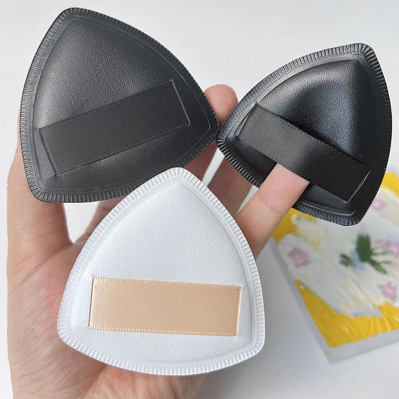 A hand holds a black triangular cosmetic foundation puff brush and a white puff brush.