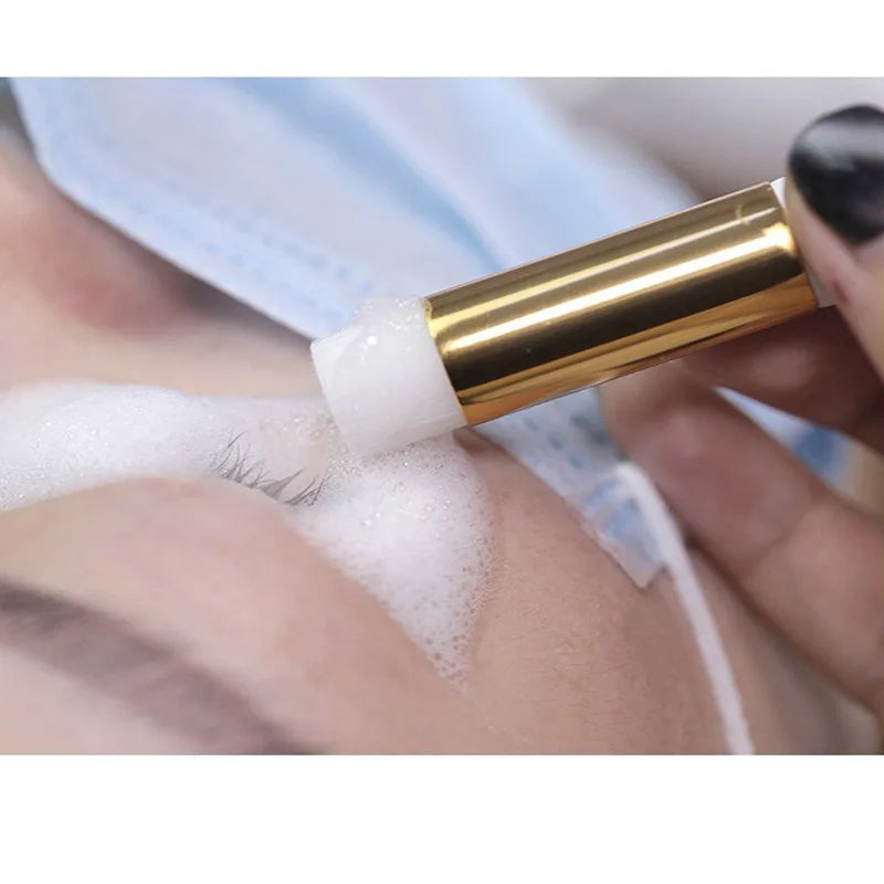 A hand is using a gold-colored bottle with a white applicator brush to clean or apply eyelash extensions.