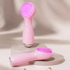 A pink vibrating LED facial cleansing brush is shown standing on a round platform.