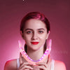 A woman is using a pink facial lifting massager with LED light therapy on her chin area.