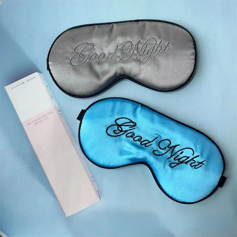 Two silk sleep masks are displayed on a light blue surface, one gray and one blue, both embroidered with Good Night.