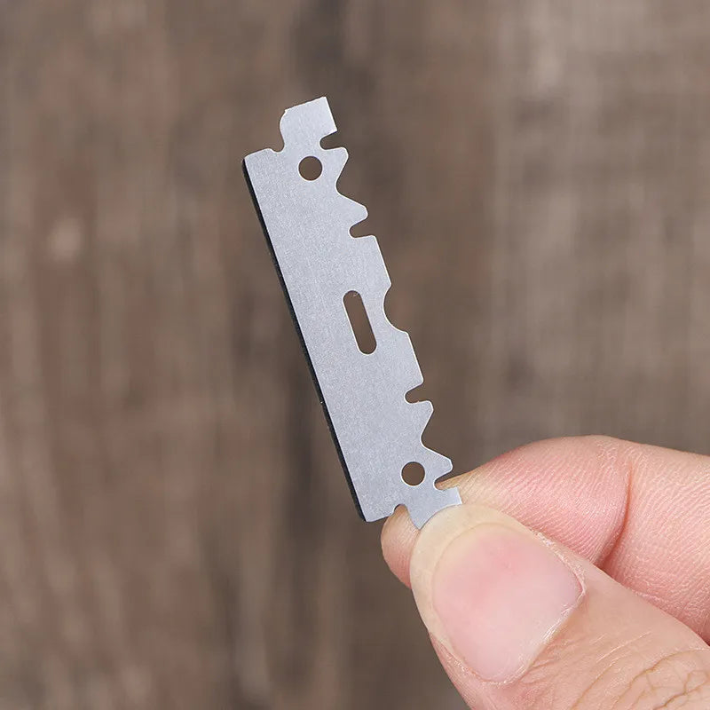 A hand holds a single stainless steel razor blade with serrated edges against a wooden background.