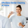 A woman is using a silver ultrasonic dental cleaning device to clean her teeth at home.
