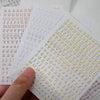 A hand holds a sheet of laser gold and silver 3D alphabet nail art stickers featuring various letters.