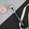 A flat top kabuki foundation brush with a pink-toned bristles rests on a black surface.