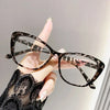 A hand is holding a pair of black and white patterned cat eye reading glasses against a light gray background.