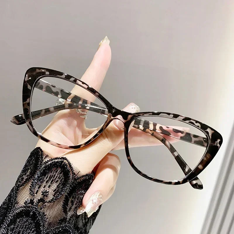 A hand is holding a pair of black and white patterned cat eye reading glasses against a light gray background.