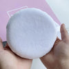 A hand holds a large, round white powder puff for makeup application against a light pink background.