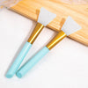 A pair of silicone facial mask applicator brushes with gold handles and light blue silicone tips resting on a wooden cutting 