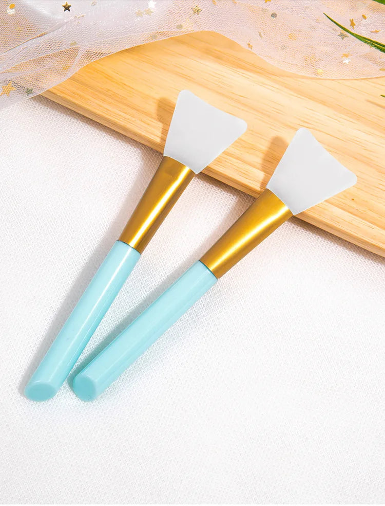 A pair of silicone facial mask applicator brushes with gold handles and light blue silicone tips resting on a wooden cutting 