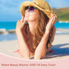 A young woman with long hair wearing a straw hat and sunglasses is relaxing on a beach, smiling.