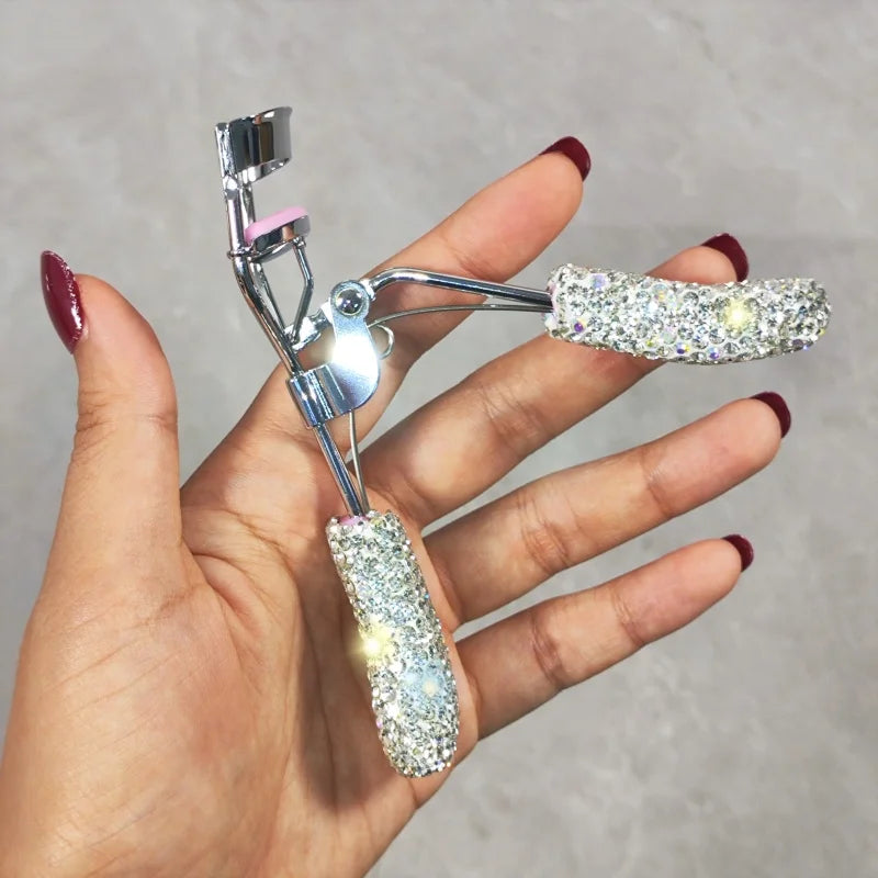 A hand holds a professional eyelash curler set with silver metal components and sparkling clear plastic curlers.