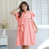 A woman wearing a pink microfiber bathrobe with a smiling face embroidery standing in front of a window.