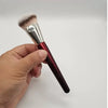 Une main tient un pinceau de maquillage liquide angulé avec une ferule argentée et une poignée noire sur fond blanc.