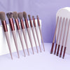 A collection of thirteen professional makeup brushes with synthetic bristles displayed on a light purple surface.