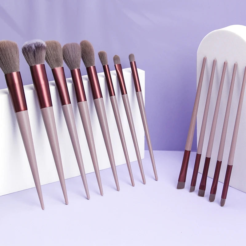 A collection of thirteen professional makeup brushes with synthetic bristles displayed on a light purple surface.