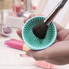 A hand holds a teal silicone makeup brush cleaner with a black brush and pink cosmetic tubes.