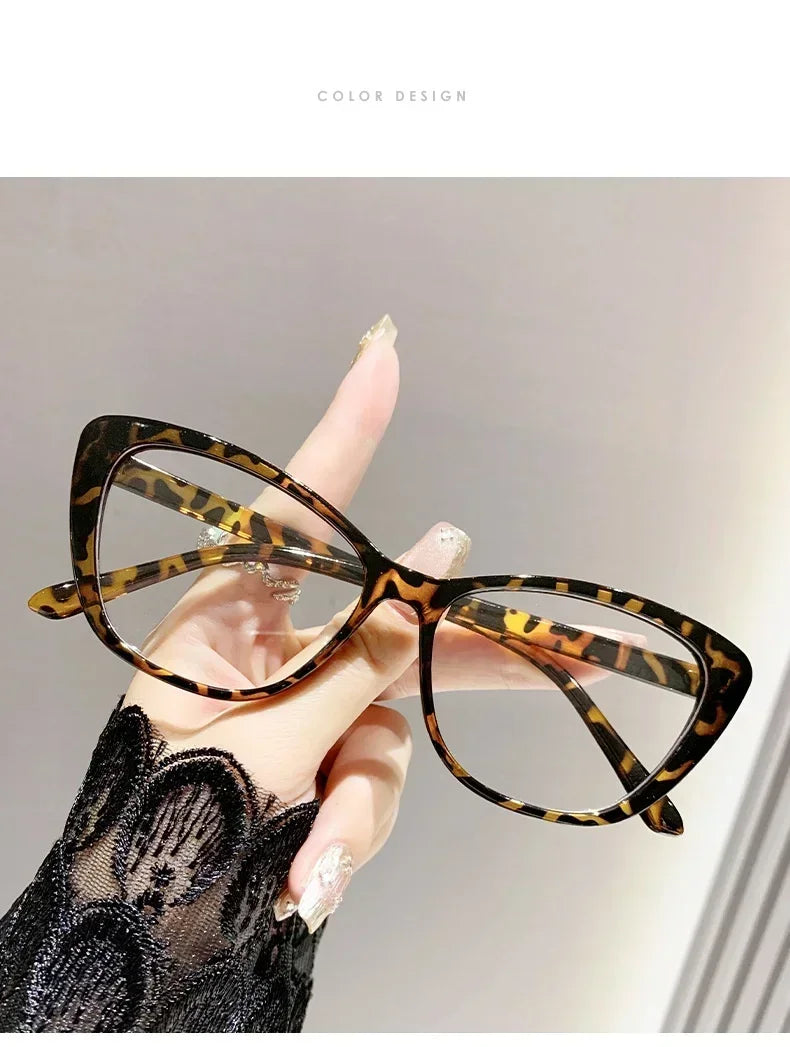 A hand is holding a pair of black and yellow patterned cat eye reading glasses against a light gray background.