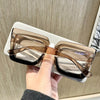 A hand holds a pair of classic square eyeglasses with clear lenses and brown frames against a light background.