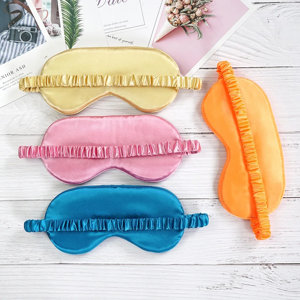 Three silk sleep eye masks in gold, pink, and blue with fringed edges displayed on a white wooden surface.