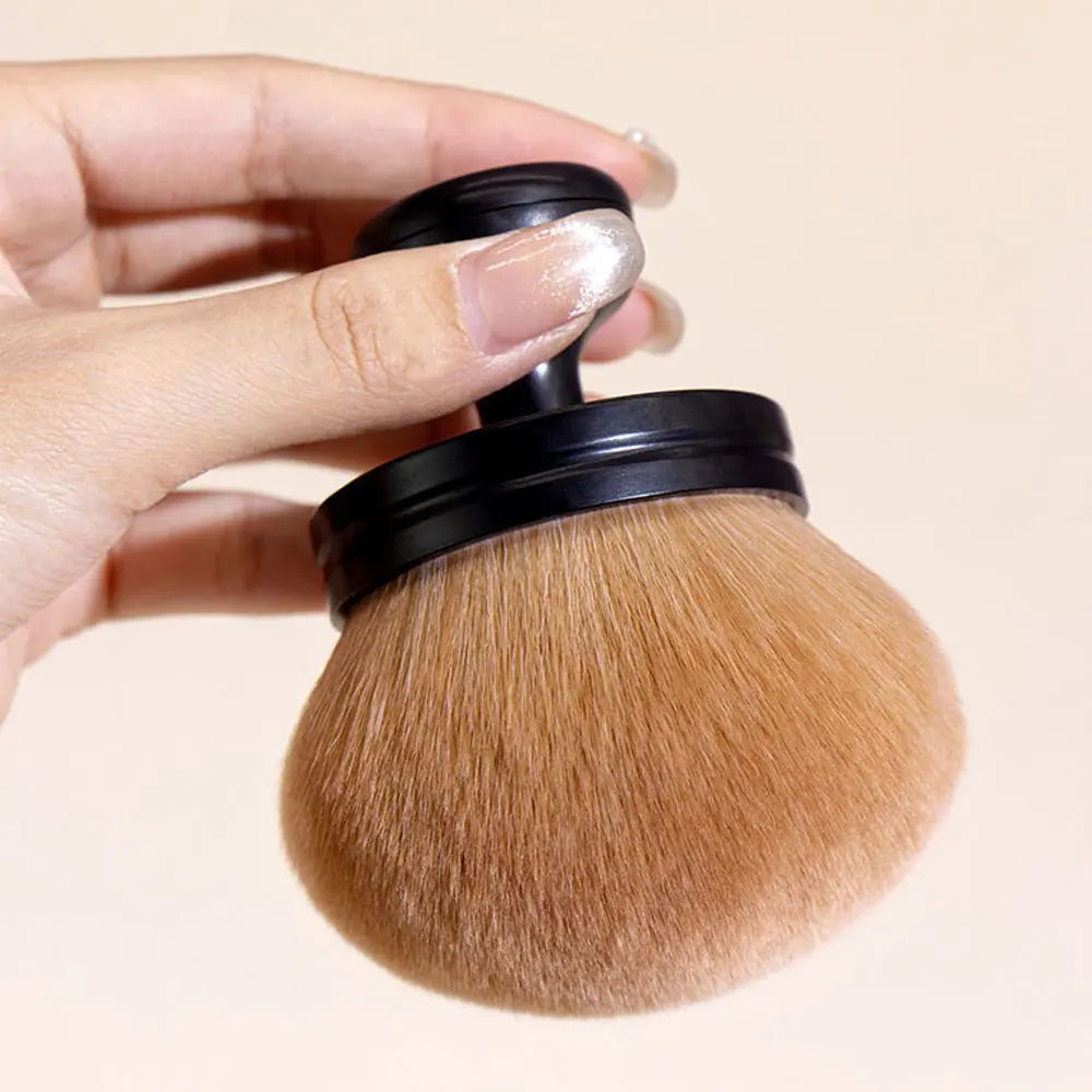 Une main tient un grand pinceau de maquillage avec une poignée noire et des poils blond doré.