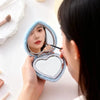 A woman holds a heart-shaped foldable travel makeup mirror with magnification, showing her reflection.