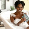 A woman is relaxing in a bathtub filled with foam, using an African net exfoliating loofah sponge.