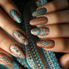 A close-up of fingers displaying 24 press on nails featuring pink and teal floral patterns on a blue fabric background.