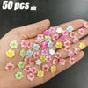 A hand holds a mix of 50 floral resin nail charms in various shades of pink, purple, and yellow.