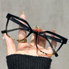Una mano sostiene unas gafas de lectura cuadradas negras anti-luz azul con detalles dorados sobre un fondo gris.