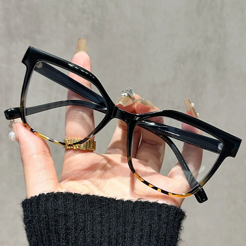 Una mano sostiene unas gafas de lectura cuadradas negras anti-luz azul con detalles dorados sobre un fondo gris.