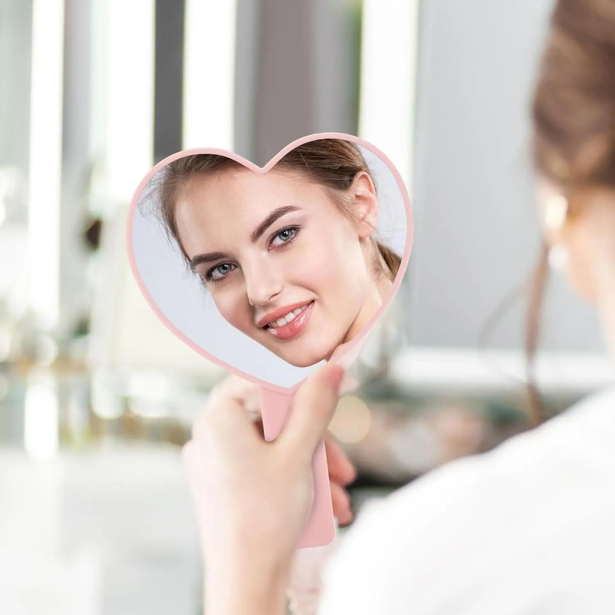 Une femme tient un miroir à cils en forme de cœur rose devant son visage.