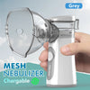 A hand holds a grey portable rechargeable nebulizer device with a clear mask, set against a light blue background.