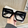 A hand holds a pair of black square eyeglasses with clear lenses against a light background.
