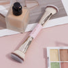 A makeup brush with a silver handle and fluffy bristles is shown next to a glass bottle of product.