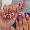 A hand displaying a set of 24-piece almond press-on nails featuring pink and white 3D floral and butterfly designs.