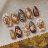 Collection de 24 ongles press-on roses à l'amande française avec paillettes dorées et accents floraux sur un tissu beige.