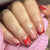 A hand displays nude pink press-on nails with leopard print accents and red 3D charms on a pink background.