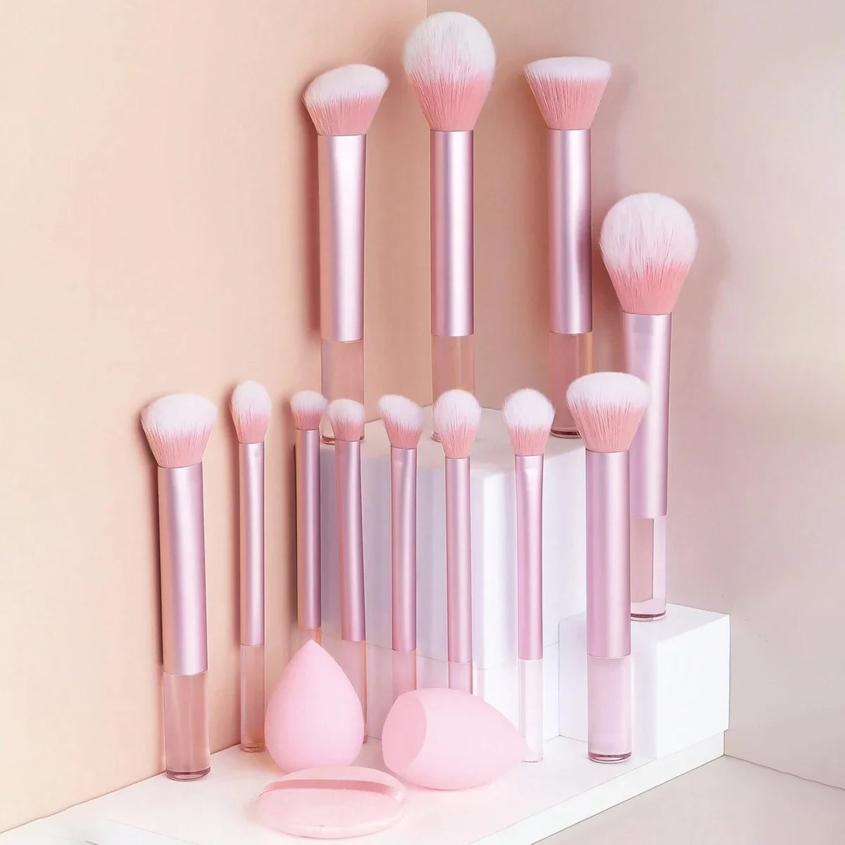 Una colección de 12 brochas de maquillaje profesionales en varios tonos rosa y nude sobre una superficie blanca.
