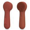 A pair of silicone facial cleansing brushes in a matte terracotta color, showing the textured brush heads.