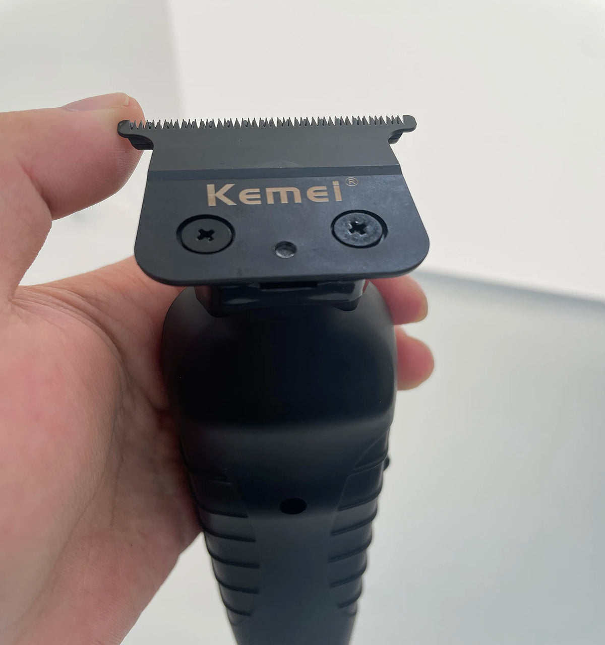 A hand holds a black electric hair clipper trimmer with a metal guard and branding visible.