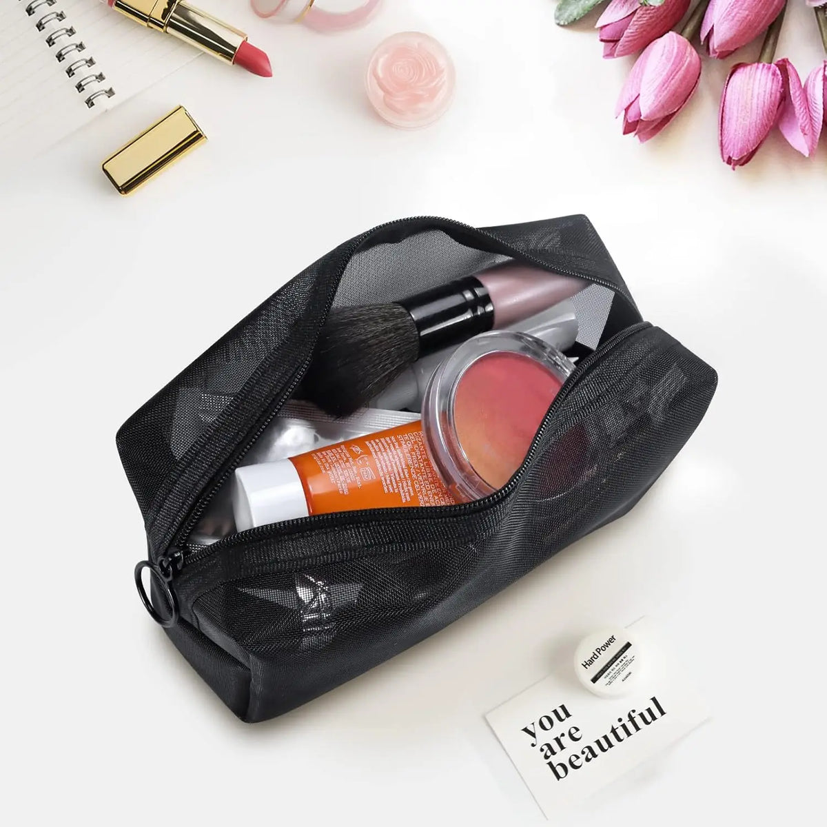 Un sac de maquillage en filet noir ouvert, montrant des pinceaux et autres produits de beauté, sur fond blanc.