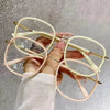 A hand holds a pair of oversized, thin-rimmed reading glasses with light pink frames and gold accents.