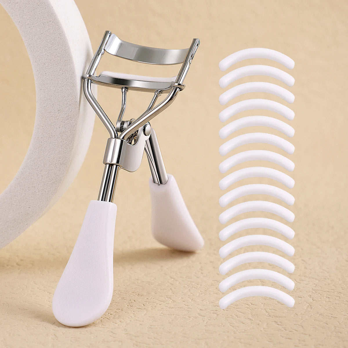 A professional stainless steel eyelash curler set with white plastic components shown on a beige background.
