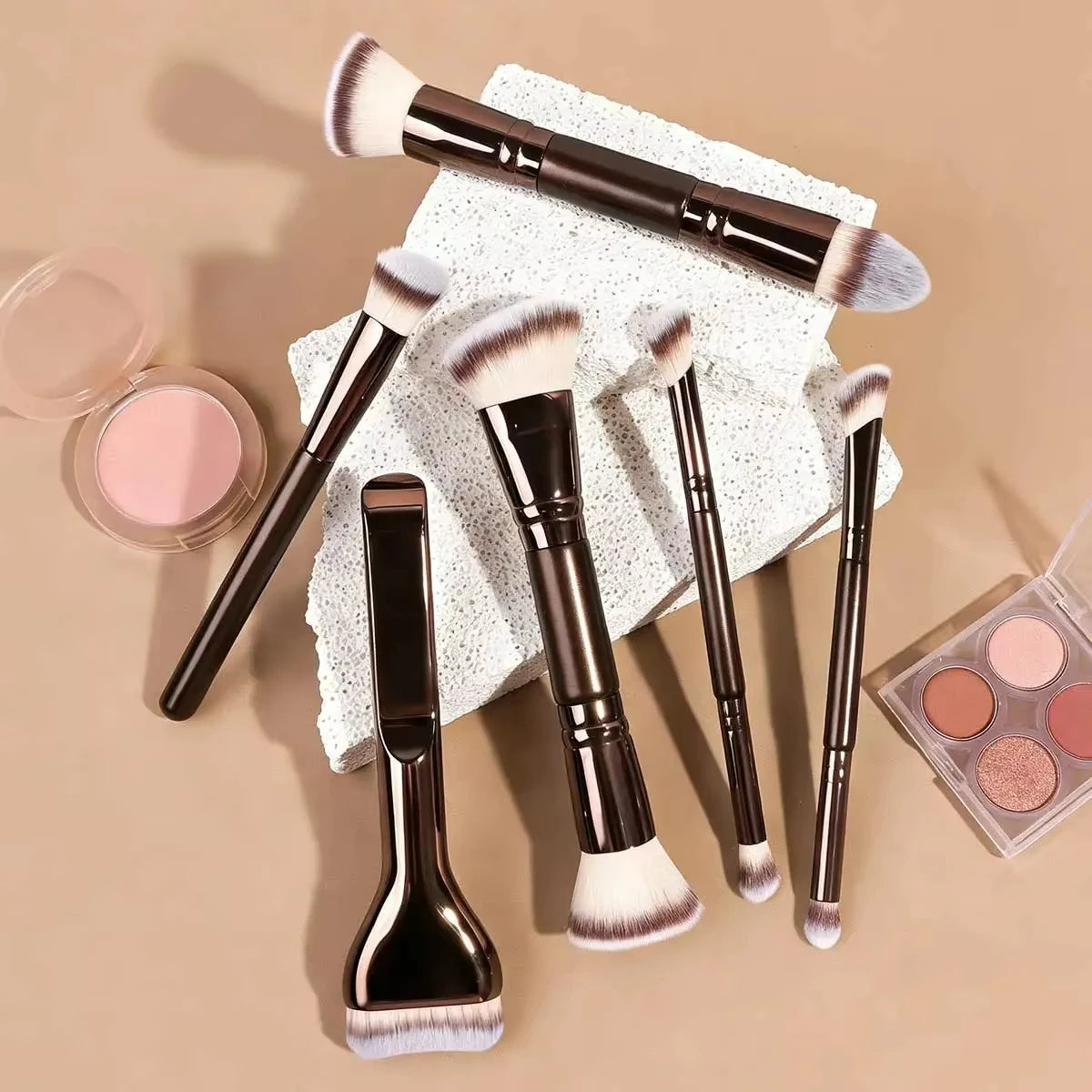 A set of seven professional makeup brushes with dark handles and light pink bristles displayed on a textured surface.